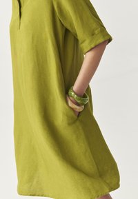 Rochie din in verde, cu o croială lejeră, mâneci scurte răsucite și buzunare laterale. Modelul poartă două brățări, una verde și una crem.