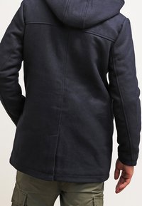Persona con giacca a cappuccio blu scuro e pantaloni cargo verde oliva, vista da dietro su uno sfondo semplice.