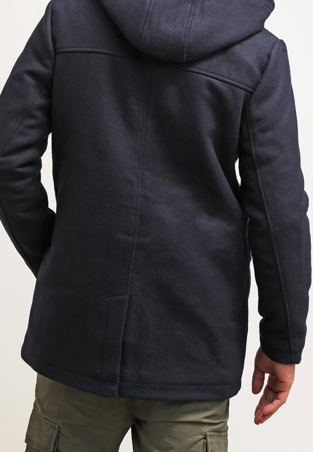 Persona con giacca a cappuccio blu scuro e pantaloni cargo verde oliva, vista da dietro su uno sfondo semplice.