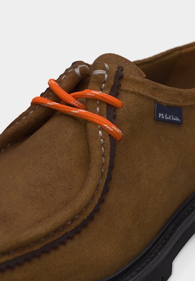 Καφέ καστόρινο παπούτσι με πορτοκαλί κορδόνια, λεπτομερείς λευκές ραφές, διακοσμητική λωρίδα με κυματιστό τελείωμα και μαύρη σόλα, που διαθέτει μικρό ύφασμα με ετικέτα της μάρκας.