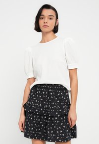 Blouse blanche texturée à manches courtes associée à une jupe noire à volants ornée d'imprimés floraux. La blouse présente un col rond et des manches bouffantes.
