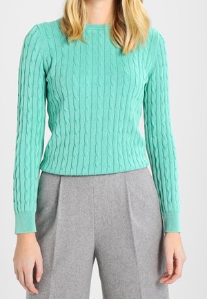 Frau trägt einen türkisfarbenen, kabelgestrickten Pullover und hoch taillierte graue Wollhosen, steht mit entspannten Armen seitlich an einem einfarbigen Hintergrund.