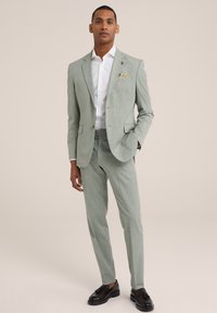 Šviesiai žalia pritaikyta švarka su subtiliu dryžuotu raštu, pasižyminti siauru siluetu ir įpjautomis apykaklėmis, derinama su balta palaidine ir tamsiais batais.