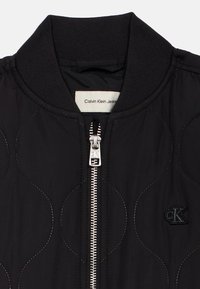 Čierna prešívaná bomber bunda s predným zipsom, ribovaným golierom a vyšitým logom na ľavej strane. Hladká textúra.