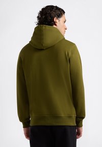 Muž s kučeravými vlasmi oblečený v olivovozelenom mikine, zobrazený zozadu na svetlom pozadí.