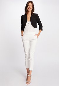 Schwarze cropped Jacke über einem weißen Hemd mit Knöpfen, kombiniert mit maßgeschneiderten weißen Hosen und silbernen, offenen Sandalen. Glatter Stoff mit elegantem Design.