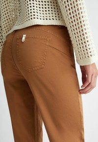 Zbliżenie na osobę w brązowych spodniach i jasnobeżowym swetrze o luźnym splotzie, ukazujące tylną kieszeń i część prawej dłoni.