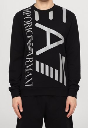Person trägt schwarzen Emporio Armani Pullover mit langen Ärmeln und großen silbernen geometrischen Formen sowie dem Buchstaben "A" auf der Vorderseite.