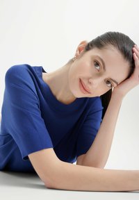 Maglione blu a maniche corte con una texture morbida e scollatura arrotondata, caratterizzato da polsini a costine. La modella poggia la testa su una mano.