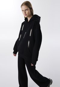 Sudadera con capucha negra y detalles en metal plateado, con un bolsillo frontal, combinada con pantalones holgados negros, contra un fondo liso.