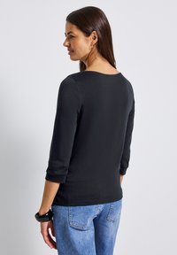 Schwarzes Oberteil mit drei Viertel-Ärmeln und rundem Ausschnitt, aus glattem Stoff, kombiniert mit hellblauen Jeans. Schlichtes Design, ohne Muster.