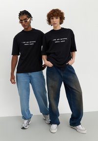 Black oversized t-shirts with white text, paired with light blue and dark blue baggy jeans. Both wear sneakers; one has sunglasses.