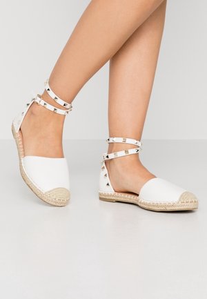 Jambes portant des espadrilles blanches plates avec des semelles en jute et des brides autour de la cheville ornées de clous pyramidaux dorés.