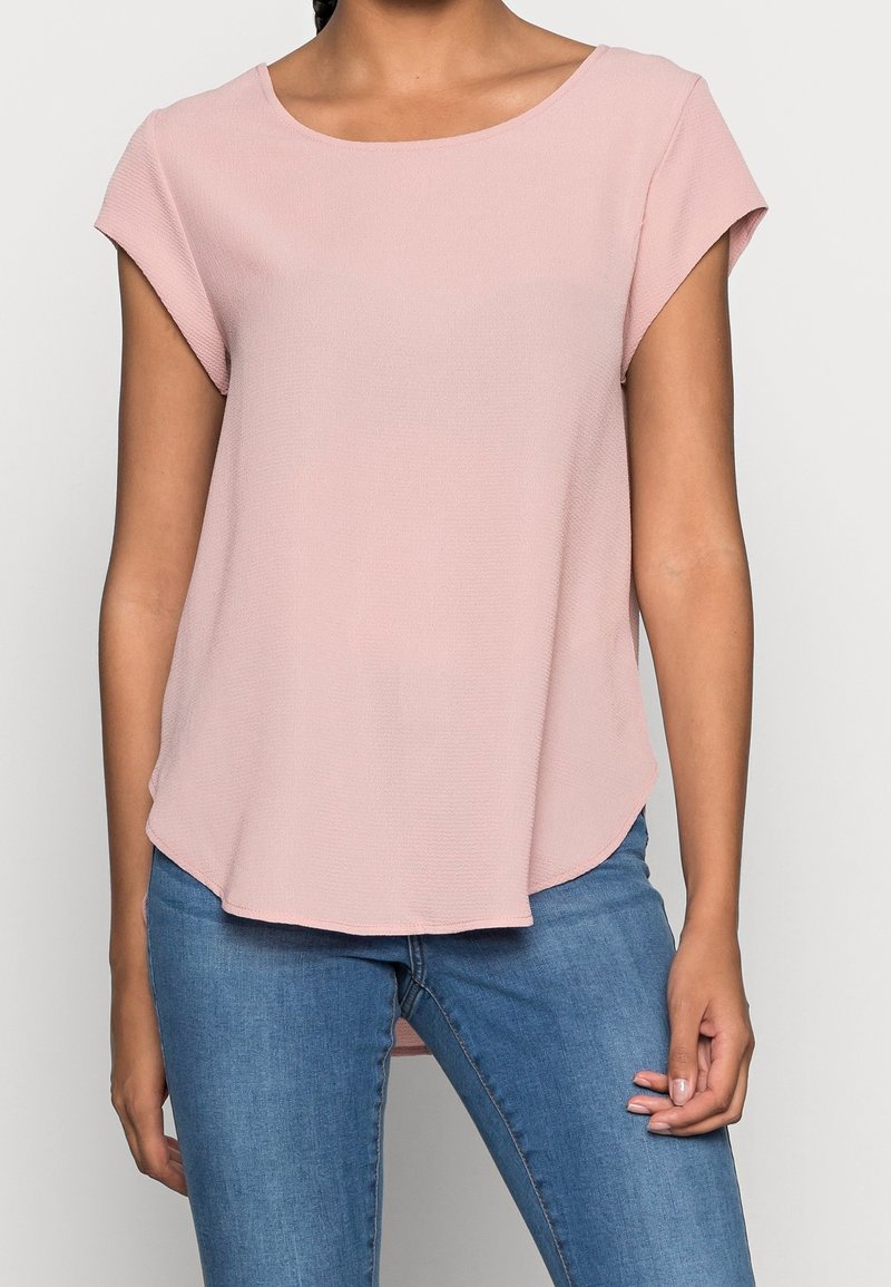 Maglietta corta a maniche corte di color rosa chiaro con scollo rotondo, tessuto fluido e orlo leggermente curvo, abbinata a jeans blu.