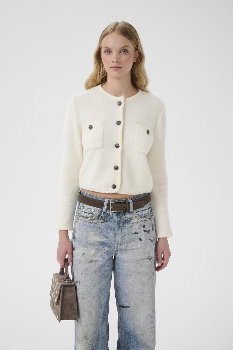 Kremo spalvos trumpas megztinis su dviem priekinėmis kišenėmis ir tamsiais mygtukais, derinamas su nusidėvėjusiais mėlynais plačios kojų dalies džinsais ir ruda rankine.