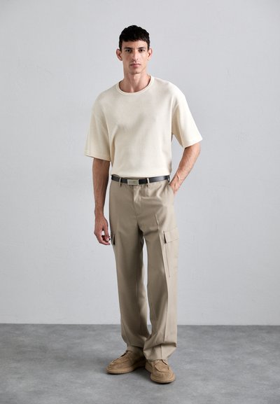 Beżowe spodnie w stylu cargo z bocznymi kieszeniami, w połączeniu z jasną, kremową, teksturowaną koszulą z krótkim rękawem i brązowymi zamszowymi butami.