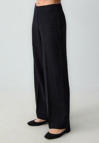 Zwarte linnen broek met wijde pijpen met een soepele textuur, hoge taille en zijrits. Geaccentueerd door een strakke, minimalistische uitstraling.