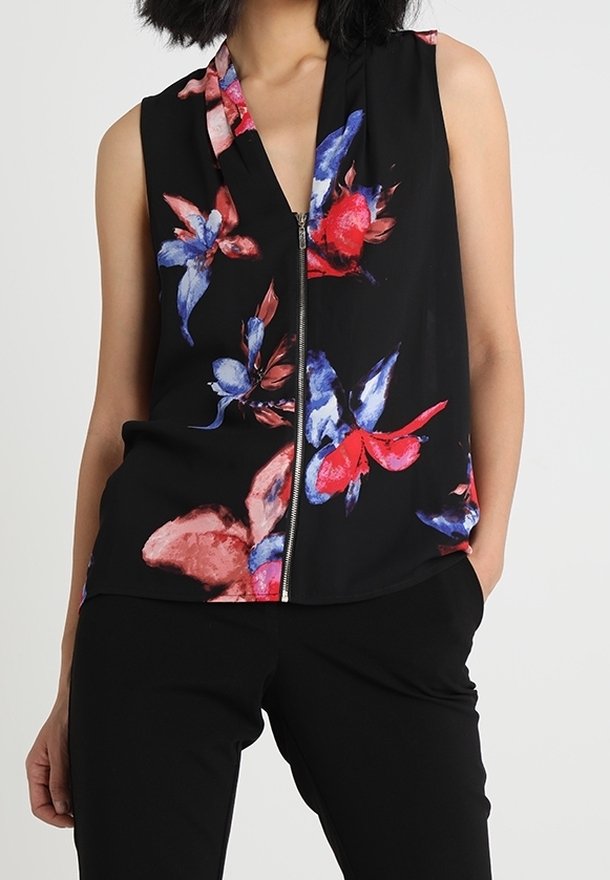 Blouse noire sans manches avec fermeture éclair à l'avant et grand motif floral rouge, bleu et rose, portée avec un pantalon noir.
