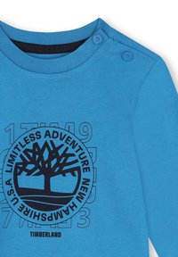 Blauwe katoenen overhemd met lange mouwen, voorzien van een zwart rond logo met een boomafbeelding en tekst. Vier schouderdrukkers voor gemakkelijk aan- en uittrekken.