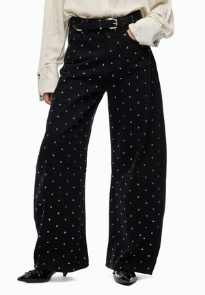 Pantaloni neri a gamba larga con borchie argento indossati con scarpe nere a punta e top beige a maniche lunghe, mano in tasca, cintura nera visibile.
