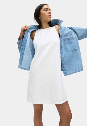 Biała sukienka bez rękawów o prostym kroju, nałożona pod jasnoniebieską kurtkę jeansową z przednią kieszenią i srebrnymi guzikami.