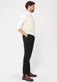 Kremowy dzianinowy bezrękawnik, biała koszula, czarne spodnie i brązowe skórzane botki za kostkę. Stylizacja charakteryzuje się luźnym krojem i gładką fakturą.