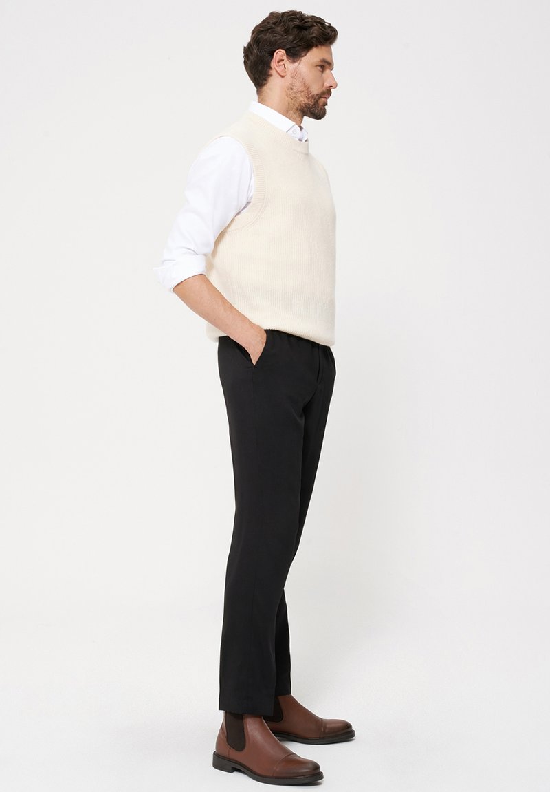 Kremowy dzianinowy bezrękawnik, biała koszula, czarne spodnie i brązowe skórzane botki za kostkę. Stylizacja charakteryzuje się luźnym krojem i gładką fakturą.