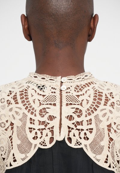 Top de renda em bege com padrões florais intricados, apresentando botões no pescoço e uma parte de trás transparente, contrastando com um tecido escuro abaixo.