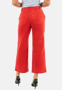 Pantalons en velours côtelé rouge à jambes larges avec deux poches arrière, une large ceinture et des détails de couture visibles, portés avec des chaussures à talons hauts noires.