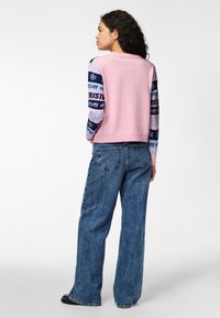 Pull en tricot rose avec des manches bleues à motifs, comportant des flocons de neige et du texte. Associé à un jean en denim bleu à coupe décontractée.