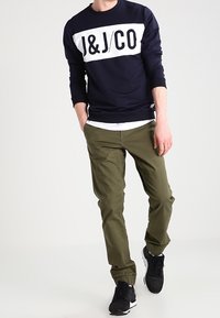 Marinblå sweatshirt med vit "J&J/CO" tryck, kombinerad med olivgröna tapered-byxor och svarta sportskor. Avslappnad, bekväm passform.