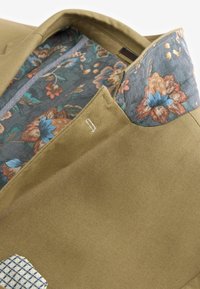 Oliven grøn blazer med et tekstureret stof. Indvendig foring har blomster mønstre i blå og orange på en mørk baggrund. Subtil syning detalje.