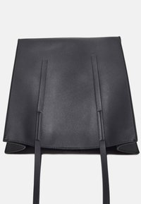 Svart läderryggsäck med ett kvadratiskt lock och två tunna axelremmar liggande platt på en vit yta.