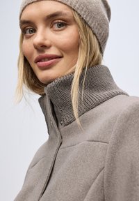 Giacca grigia chiara in misto lana con colletto alto a costine e chiusura a pressione; presenta una texture liscia e dettagli di cucitura sottili.