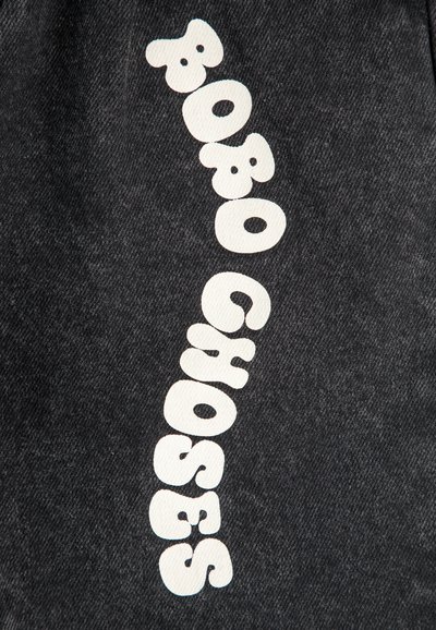 Tela de mezclilla negra con grandes letras blancas juguetonas que deletrean "BOBO CHOSES" en diagonal. La textura es ligeramente áspera y sin adornos.
