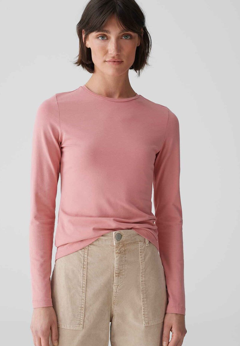 Langärmliges rosa Oberteil aus Baumwolle, mit Rundhalsausschnitt und taillierter Silhouette, kombiniert mit beigen Cordhosen.