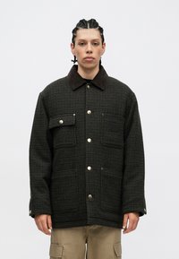 Chaqueta a cuadros en negro y marrón con un collar marrón, botones a presión dorados y dos bolsillos frontales. Tejido texturizado con un corte relajado.