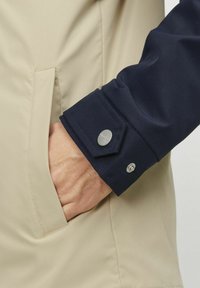 Jachetă bej cu manșetă bleumarin, având un detaliu cu buton tip snap. Materialul pare neted și ușor, cu un design curat și buzunar ascuns.
