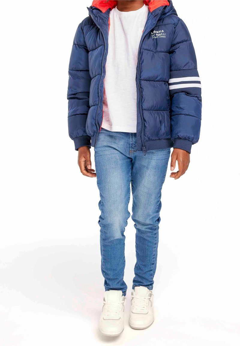 Garçon portant une doudoune bleu marine avec doublure rouge, un t-shirt blanc, un jean bleu clair et des baskets blanches, debout devant un fond blanc.