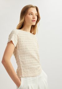 Top corto di un beige chiaro con texture, presenta un motivo a pieghe. Abbinato a pantaloni bianchi plissettati. Sfondo semplice.