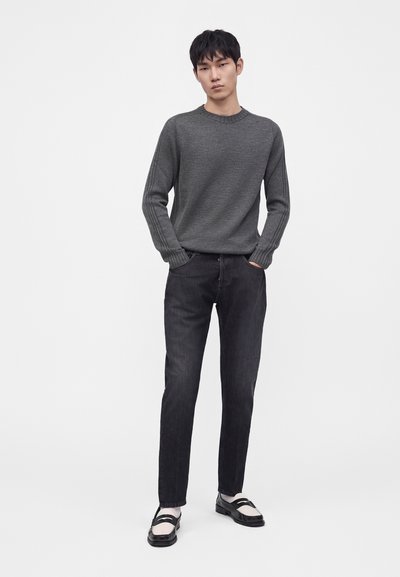 Jeune homme debout les mains dans les poches, portant un pull gris foncé, un jean noir, des chaussettes blanches et des mocassins noir et blanc sur fond blanc uni.