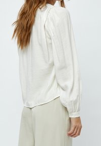 Bílá halenka s dlouhými rukávy, sbíranými rameny a knoflíkovými manžetami, vyrobená z měkké texturované látky a s uvolněným střihem.