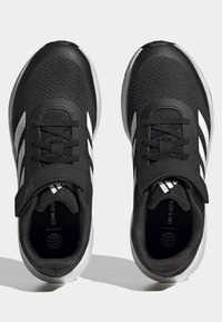 Baskets de sport noires avec tissu en maille, rayures blanches, fermetures à lacets et Velcro, bout arrondi et semelle en caoutchouc. Design simple et palette de couleurs neutres.