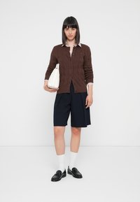Brun kabelstrikket cardigan over en lyseblå skjorte med krage, kombinert med marineblå shorts, hvite sokker og svarte loafers. Modellen holder en hvit veske.
