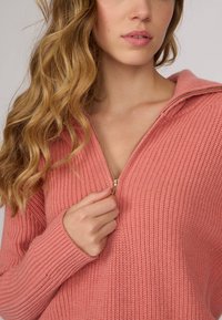 Pull à fermeture éclair tricoté en rose doux. Présente une texture côtelée, des manches longues et un détail de fermeture éclair doré au niveau du col.