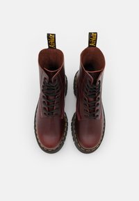 Botas de tobillo de cuero burdeos con punta redonda, cordones negros, suela negra y costuras amarillas. Incluyen un tirador negro.