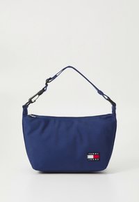 Skuldertaske i marineblåt stof med lynlås, der har et lille logo-patch i rødt, hvidt og blåt, justerbar rem og blød tekstur.