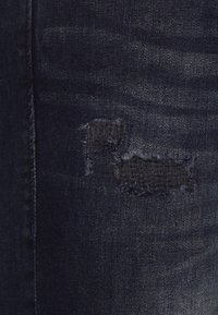 Mörkblå denimjeans med ett slitna lappar, som har två små fransiga hål. Tyget har en slät textur med subtil blekning.