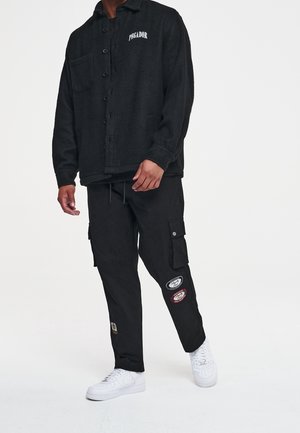 Sort uld knapjakke med en brystlomme og logo. Sorte cargo bukser med broderede lapper. Hvide sneakers.
