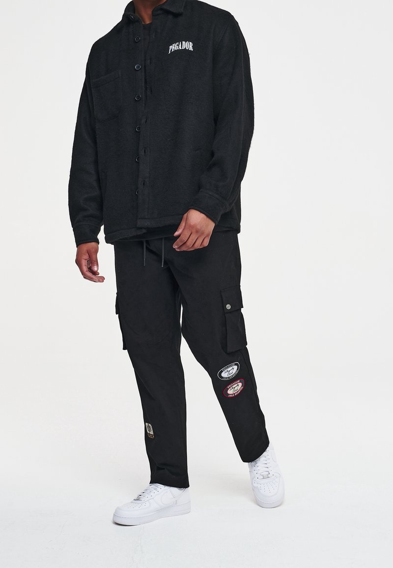 Chaqueta de lana negra con botones, bolsillo en el pecho y logotipo. Pantalones cargo negros con parches bordados. Zapatillas blancas.